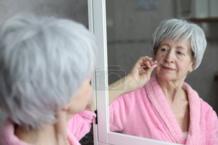 Photo for Close-up portrait of mature woman pulling out facial hair in front of mirror in bathroom - Royalty Free Image