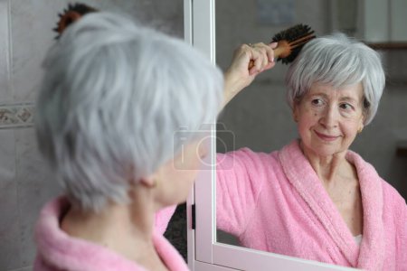 Photo for Close-up portrait of mature woman brushing hair in front of mirror in bathroom - Royalty Free Image