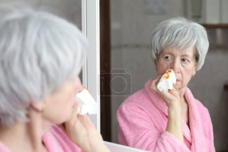 Foto de Retrato de cerca de la mujer madura con la nariz sangrante delante del espejo en el baño - Imagen libre de derechos