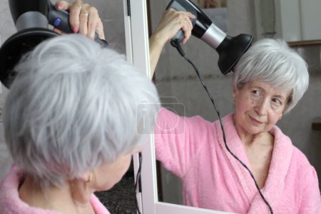 Photo for Close-up portrait of mature woman with hair dryer in front of mirror in bathroom - Royalty Free Image