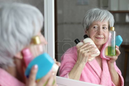 Foto de Primer plano retrato de mujer madura con botellas de cuidado de la piel delante del espejo en el baño - Imagen libre de derechos