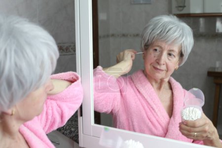 Photo for Close-up portrait of mature woman with cotton sticks brushing ears in front of mirror in bathroom - Royalty Free Image