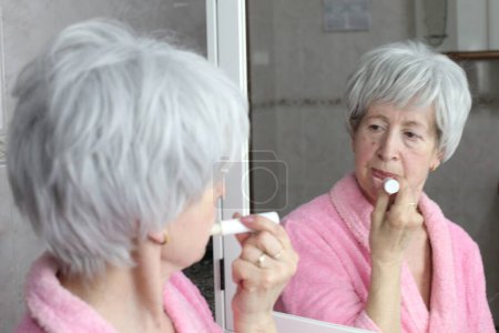 Téléchargez les photos : Portrait en gros plan de femme mature appliquant baume à lèvres devant le miroir dans la salle de bain - en image libre de droit