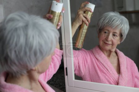Téléchargez les photos : Portrait en gros plan de femme mature appliquant de la laque à cheveux devant le miroir dans la salle de bain - en image libre de droit