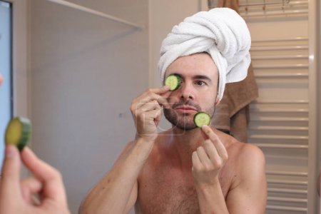 Photo for Portrait of young man with towel on head and cucumbers on eyes in front of mirror in bathroom - Royalty Free Image