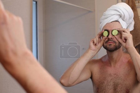 Téléchargez les photos : Portrait de jeune homme avec serviette sur la tête et concombres sur les yeux en face du miroir dans la salle de bain - en image libre de droit