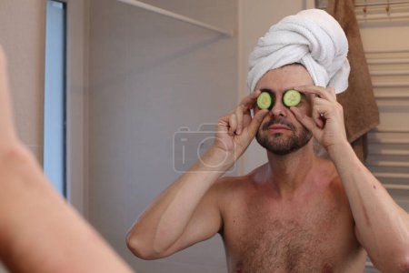 Téléchargez les photos : Portrait de jeune homme avec serviette sur la tête et concombres sur les yeux en face du miroir dans la salle de bain - en image libre de droit