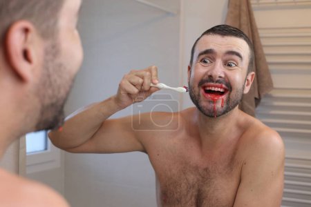 Téléchargez les photos : Portrait de jeune homme avec des gencives saignantes se brossant les dents devant le miroir dans la salle de bain - en image libre de droit