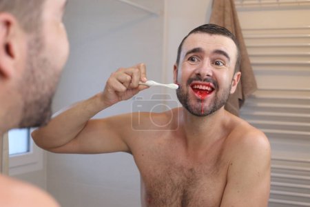Photo for Portrait of young man with bleeding gums brushing his teeth in front of mirror in bathroom - Royalty Free Image