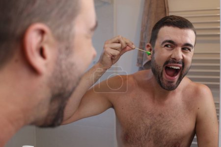Téléchargez les photos : Portrait de jeune homme épilant ses cheveux de l'oreille devant le miroir dans la salle de bain - en image libre de droit