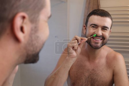Téléchargez les photos : Portrait de jeune homme épilant ses poils du nez devant le miroir dans la salle de bain - en image libre de droit