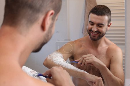Téléchargez les photos : Portrait de jeune homme rasant son bras devant le miroir dans la salle de bain - en image libre de droit