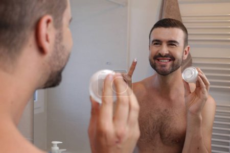 Téléchargez les photos : Portrait de jeune homme avec pot de crème hydratante devant le miroir dans la salle de bain - en image libre de droit