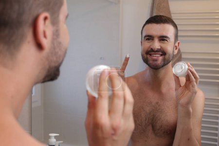 Foto de Retrato de hombre joven con frasco de crema hidratante delante del espejo en el baño - Imagen libre de derechos