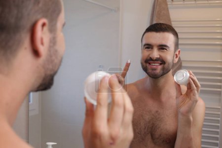 Téléchargez les photos : Portrait de jeune homme avec pot de crème hydratante devant le miroir dans la salle de bain - en image libre de droit
