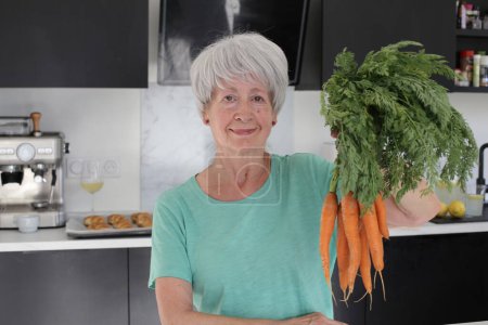 Photo for Close-up portrait of mature woman holding bunch of carrots at kitchen - Royalty Free Image