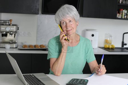 Téléchargez les photos : Portrait en gros plan de femme mature parlant par téléphone tout en travaillant avec un ordinateur portable à la cuisine - en image libre de droit