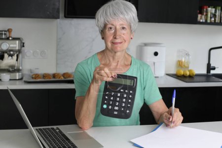 Foto de Primer plano retrato de mujer madura con ordenador portátil y calculadora contando impuestos en la cocina - Imagen libre de derechos