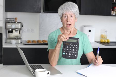 Téléchargez les photos : Portrait en gros plan de femme mature avec ordinateur portable et calculatrice comptant les taxes à la cuisine - en image libre de droit
