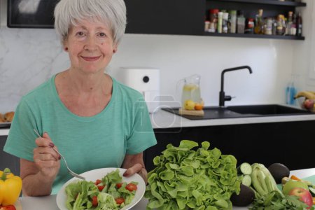 Téléchargez les photos : Portrait en gros plan d'une femme mûre mangeant une salade de légumes saine à la cuisine - en image libre de droit