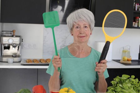 Téléchargez les photos : Portrait en gros plan d'une femme mature tenant une raquette électrique anti-insectes à la cuisine - en image libre de droit