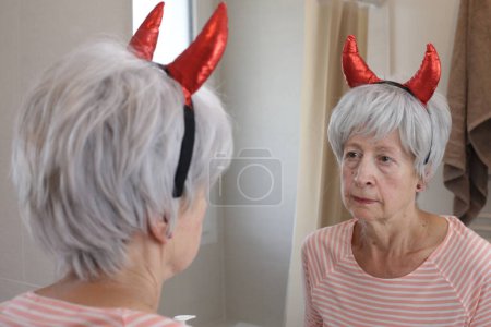 Téléchargez les photos : Portrait en gros plan de femme mature avec des cornes du diable rouge devant le miroir dans la salle de bain - en image libre de droit