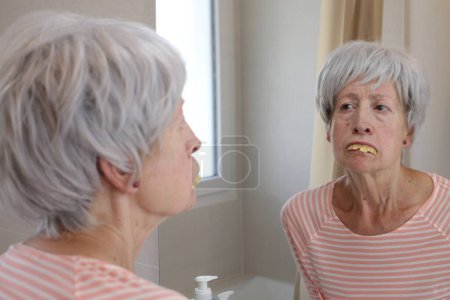 Téléchargez les photos : Portrait en gros plan de femme mature regardant ses dents devant le miroir dans la salle de bain - en image libre de droit