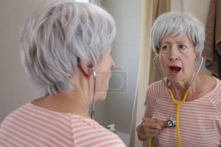 Photo for Close-up portrait of mature woman with stethoscope listening to her heart in front of mirror in bathroom - Royalty Free Image