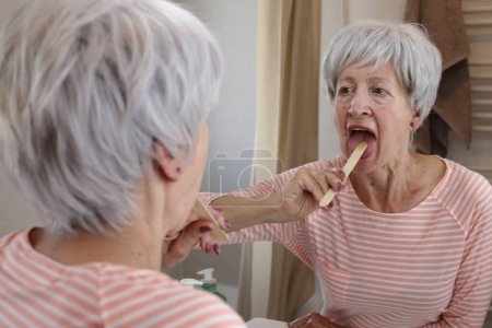 Photo for Close-up portrait of mature woman examining her mouth with wooden stick in front of mirror in bathroom - Royalty Free Image
