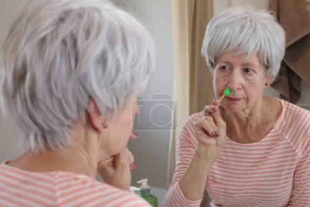 Photo for Close-up portrait of mature woman removing her nose hair with wax in front of mirror in bathroom - Royalty Free Image