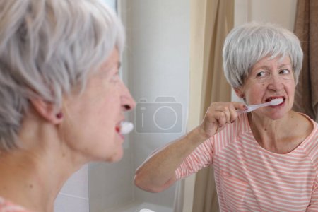 Téléchargez les photos : Portrait en gros plan de femme mature avec des gencives saignantes pendant qu'elle se brosse les dents devant le miroir dans la salle de bain - en image libre de droit