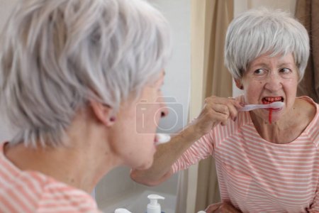 Téléchargez les photos : Portrait en gros plan de femme mature avec des gencives saignantes pendant qu'elle se brosse les dents devant le miroir dans la salle de bain - en image libre de droit