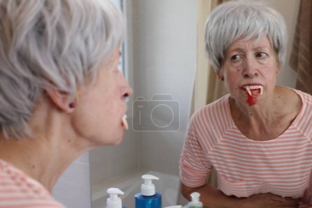 Téléchargez les photos : Portrait en gros plan de femme mature avec des dents de vampire devant le miroir dans la salle de bain - en image libre de droit