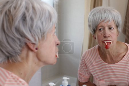 Photo for Close-up portrait of mature woman with vampire teeth in front of mirror in bathroom - Royalty Free Image