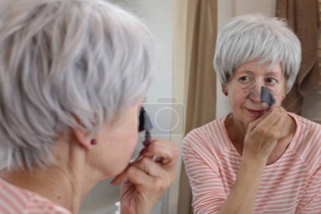 Foto de Retrato de cerca de la mujer madura con parche negro en la nariz delante del espejo en el baño - Imagen libre de derechos