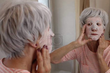 Téléchargez les photos : Portrait en gros plan de femme mature avec masque facial devant le miroir dans la salle de bain - en image libre de droit