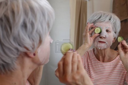 Téléchargez les photos : Portrait en gros plan de femme mature avec masque facial devant le miroir dans la salle de bain - en image libre de droit