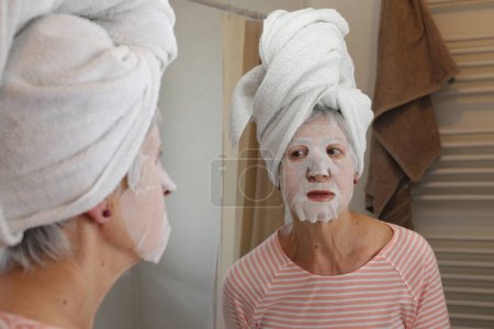Téléchargez les photos : Portrait en gros plan de femme mature avec masque facial et serviette sur la tête devant le miroir dans la salle de bain - en image libre de droit