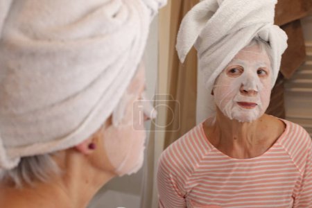 Téléchargez les photos : Portrait en gros plan de femme mature avec masque facial et serviette sur la tête devant le miroir dans la salle de bain - en image libre de droit