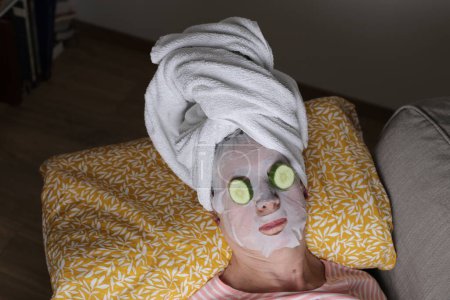 Téléchargez les photos : Portrait en gros plan de femme mature relaxante avec serviette sur la tête, masque facial et concombre sur les yeux - en image libre de droit