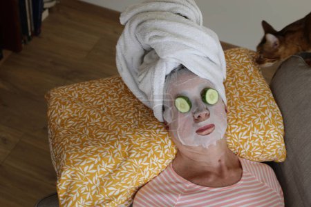 Photo for Close-up portrait of mature woman relaxing with towel on head, facial mask and cucumbers on eyes - Royalty Free Image