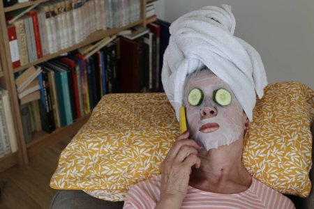 Téléchargez les photos : Portrait en gros plan de femme mature relaxante avec serviette sur la tête, masque facial et concombre sur les yeux - en image libre de droit