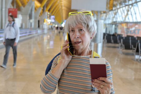 Téléchargez les photos : Portrait en gros plan de femme mature avec passeport parlant par téléphone à l'aéroport - en image libre de droit