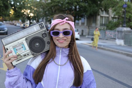 Téléchargez les photos : Portrait de la femme hipster élégant portant bandeau, lunettes de soleil et percing tenant magnétophone posant sur fond urbain - en image libre de droit