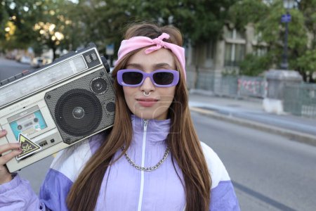 Téléchargez les photos : Portrait de la femme hipster élégant portant bandeau, lunettes de soleil et percing tenant magnétophone posant sur fond urbain - en image libre de droit