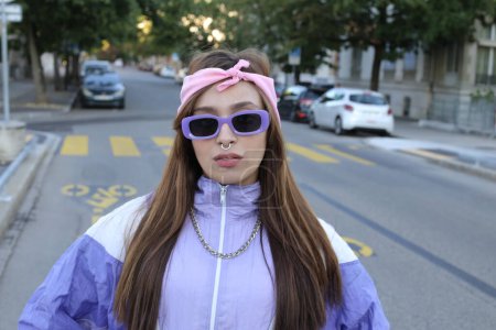 Téléchargez les photos : Portrait de femme hipster élégante portant bandeau, lunettes de soleil et piercing sur fond urbain - en image libre de droit