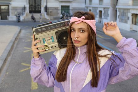 Téléchargez les photos : Élégant hipster femme portant bandeau et percing tenant magnétophone sur fond urbain - en image libre de droit