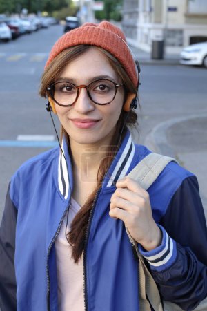 Téléchargez les photos : Étudiante portant un bonnet et des lunettes écoutant de la musique via un casque marchant à l'extérieur - en image libre de droit