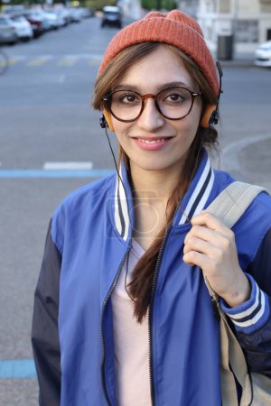 Photo for Student woman wearing beanie hat and eyeglasses listening to music via headphones walking outdoors - Royalty Free Image