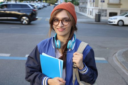 Photo for Student woman wearing beanie hat and eyeglasses holding notebooks walking outdoors - Royalty Free Image